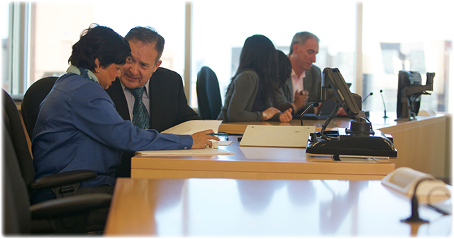 A man and woman review their paperwork at their seats in a hearing room. In the background, other people are sitting at their desks.